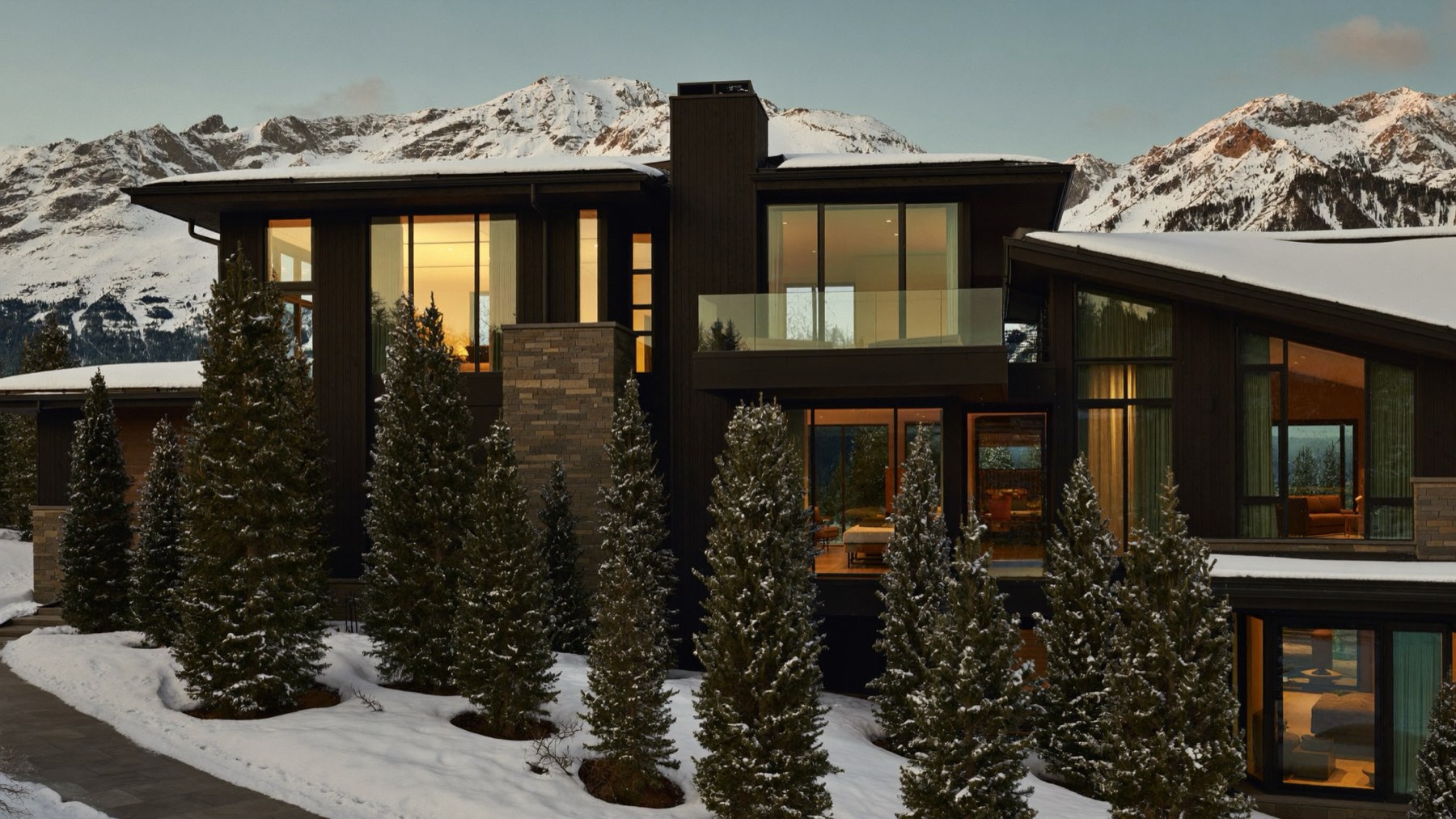 Luxury mountain home at dusk with warm light in the windows, surrounded by snow-covered pines